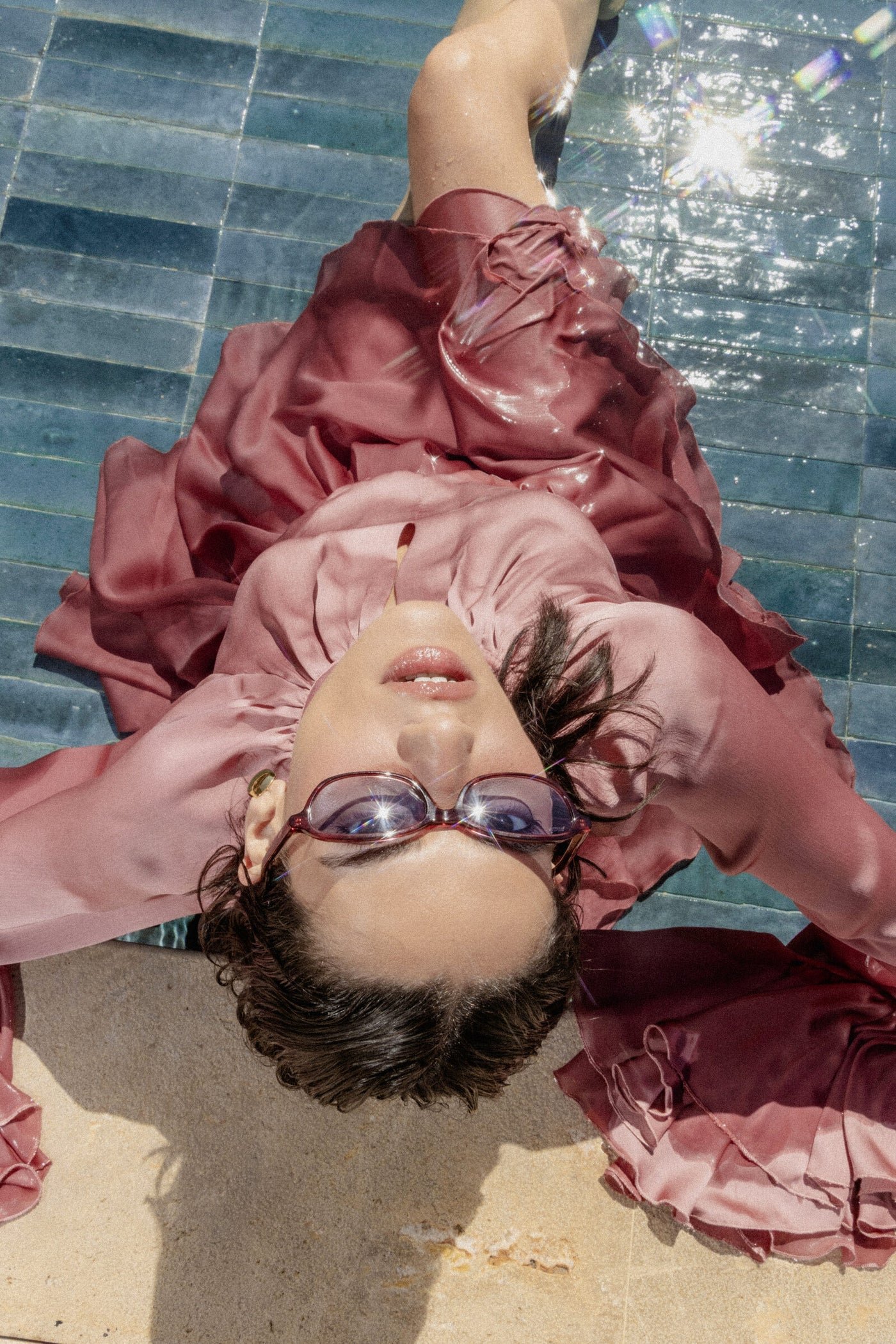 A woman in a rose dress lying at the edge of a sunlit pool, wearing mair sunglasses